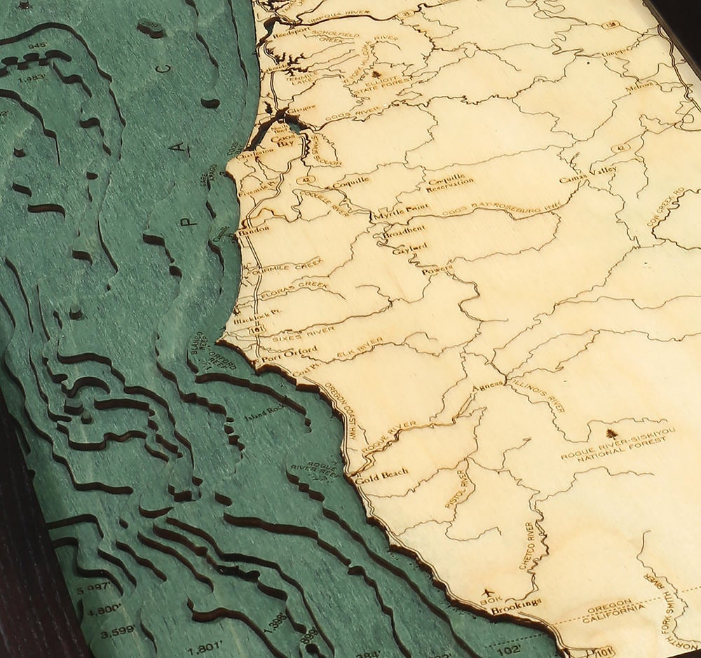 Oregon Coast 3D layered wood map in laser-cut Baltic birch, shown framed against a light wall